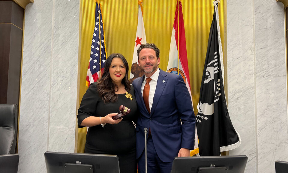 Nora Vargas and Nathan Fletcher stand in front of flags. Vargas is holding a gavel.