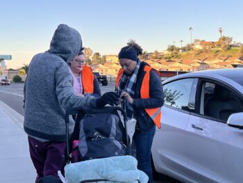 2 volunteers speak to someone wearing a sweatshirt next to a car