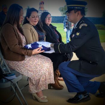Man in uniform hands folded flag to woman sitting in chair.