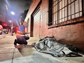 Outreach worker in orange vest kneels next to man sleeping on sidewalk