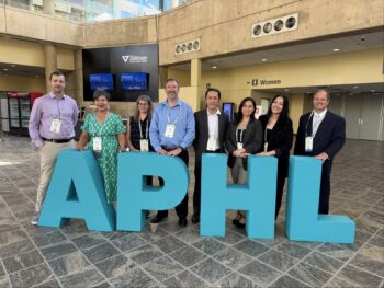 8 people posing with large letters APHL
