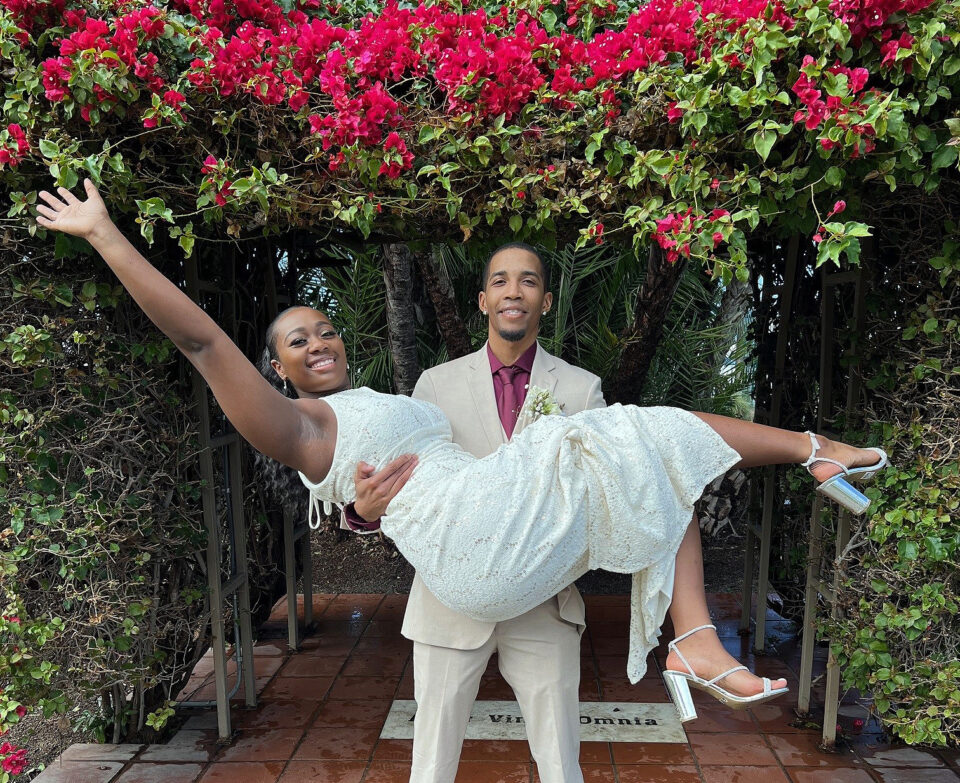 Groom holds bride under a arch of vines and flowers outside