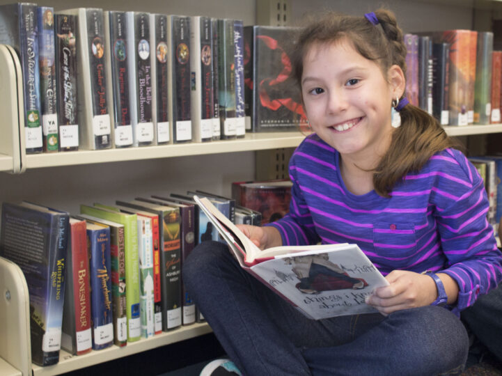 girl in purple top reading a book with bookshelf in background