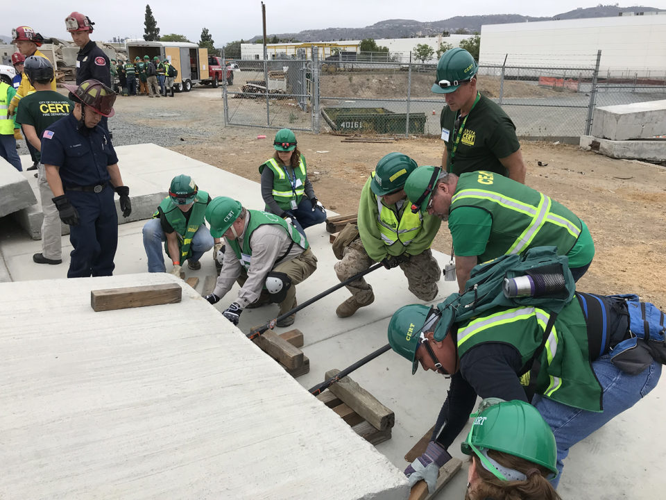 People in green vests, green helmets try to lift a heavy piece of concrete
