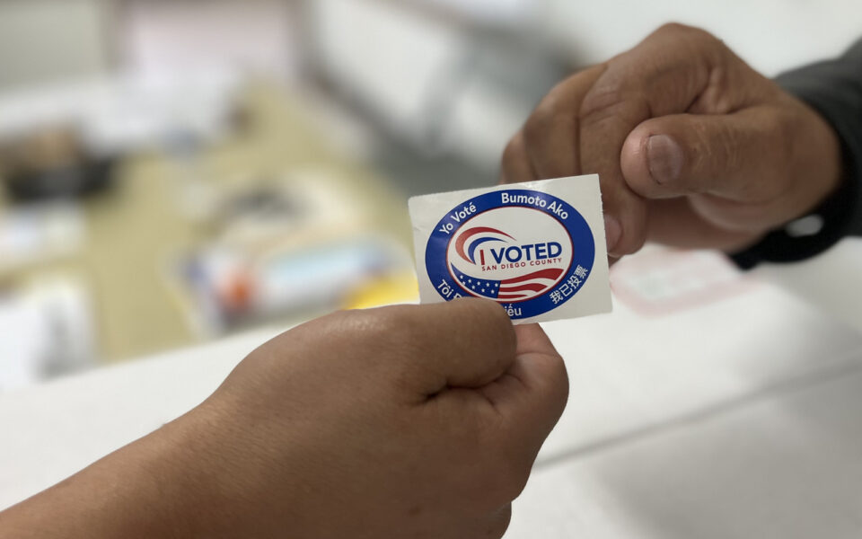 person hands "I voted" sticker over to voter