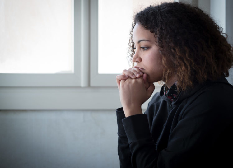 A woman sits in front of a window with her hands folded