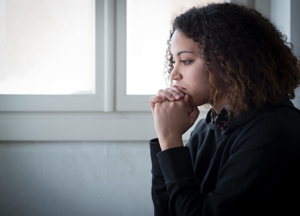 A woman sits in front of a window with her hands folded