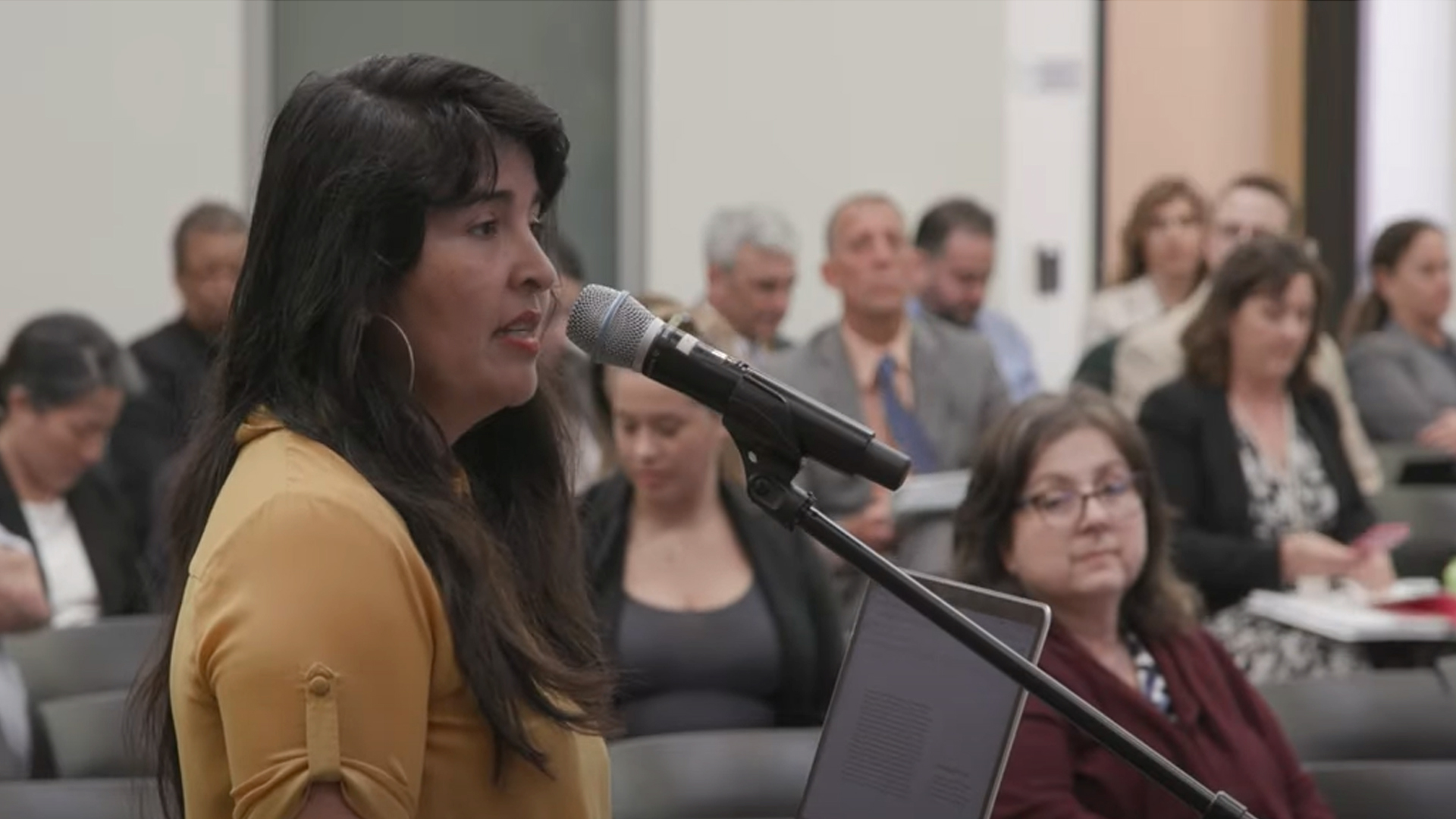 person stands in front of seat crowd and speaks at microphone