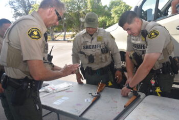 3 deputies at a table, 1 deputy puts a safety on a rifle