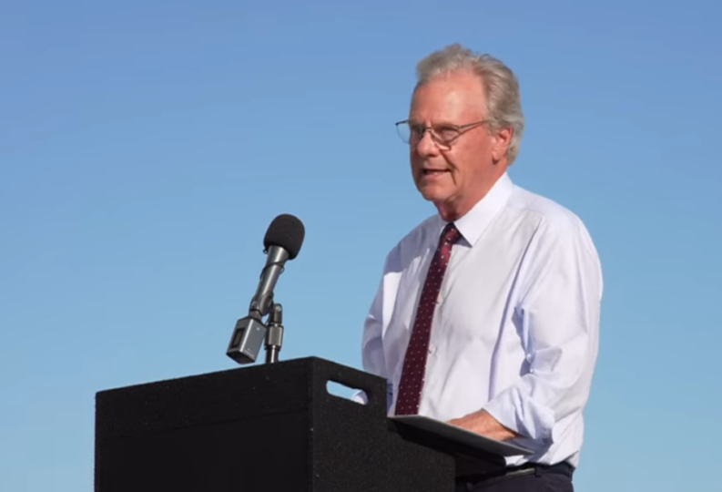 County Supervisor Jim Desmond wearing a white shirt and tie speaking at a podium in front of a blue sky.