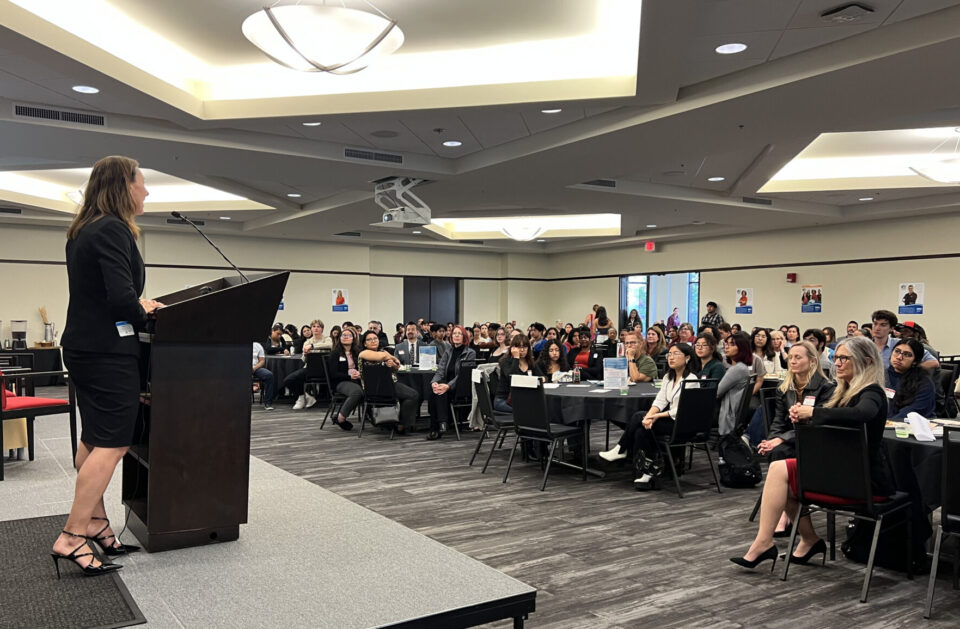 person stands at podium on stage and speaks to a room full of people at big tables
