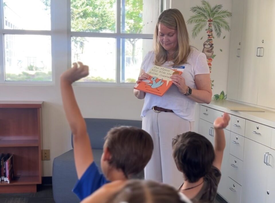 Librarian holding book leading reading vircle and the back of two kids raising their hands