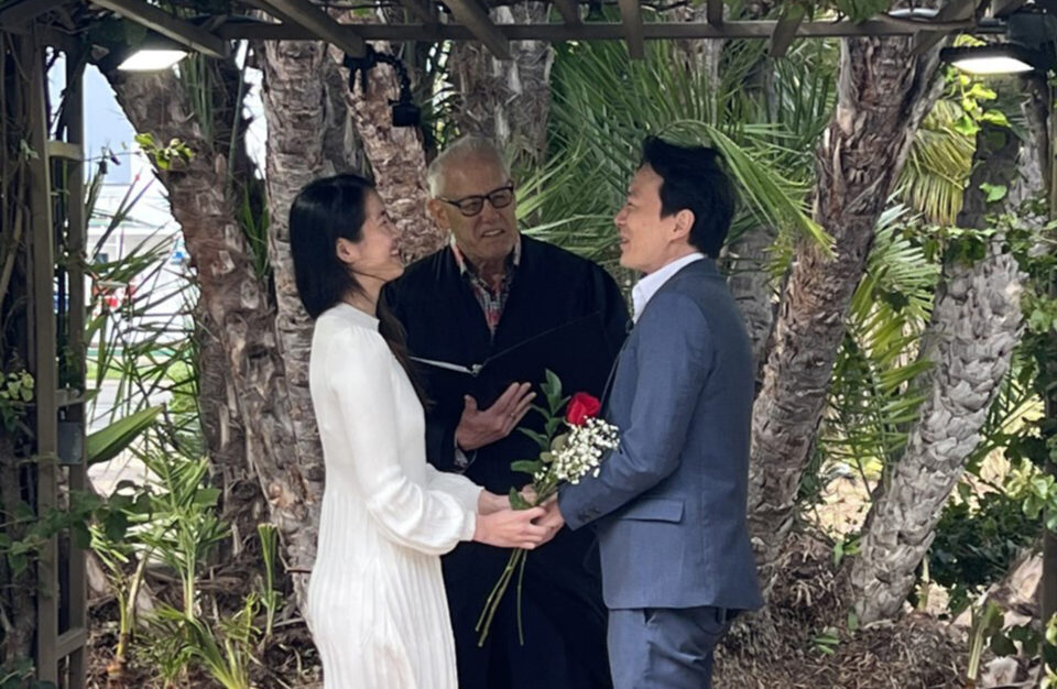 couple stand in wedding arch holding hands as someone in black rope officiates