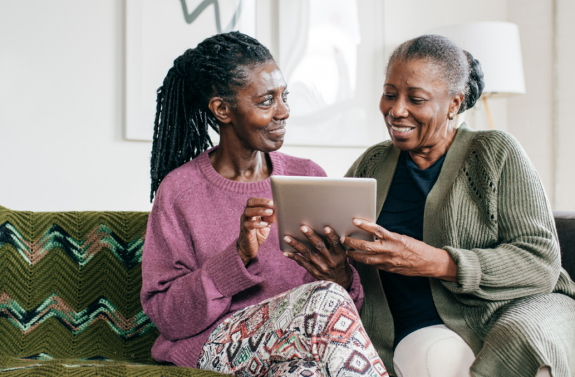 Two women, one younger and one an older adult, sit on a couch and interact while holding a tablet