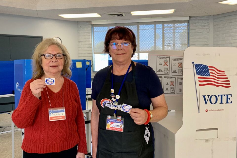 2 election workers stand in polling place holding stickers