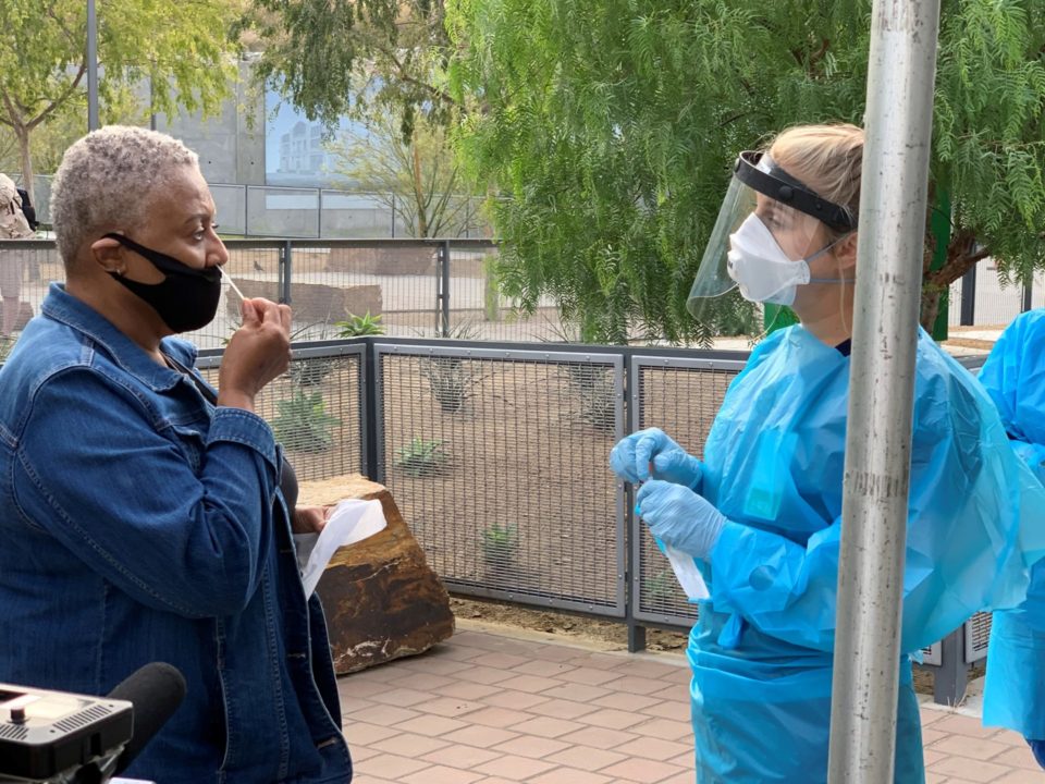 Woman gives herself a COVID-19 test and a nurse stands across from her