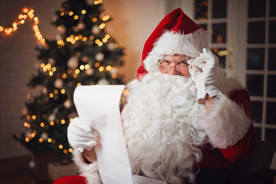 Santa with hand up to glasses and other hand holding a long list