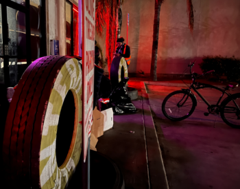Volunteer stands in the background with a large tire in the front. Volunteer speaks with person experiencing homelessness.