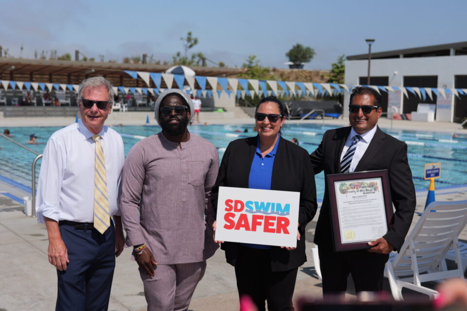4 people stand in front of pool. 1 person holds sign that says "SD Swim Safer"
