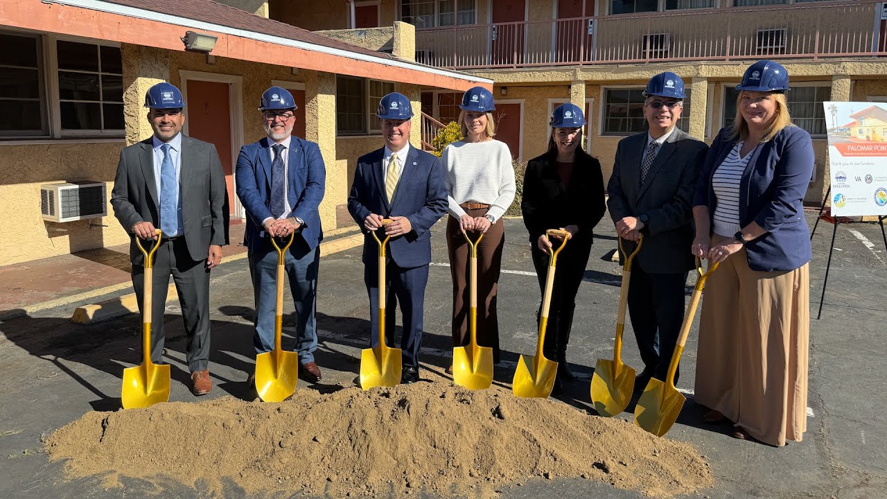 7 people stand with shovels and dirt in front of bulding