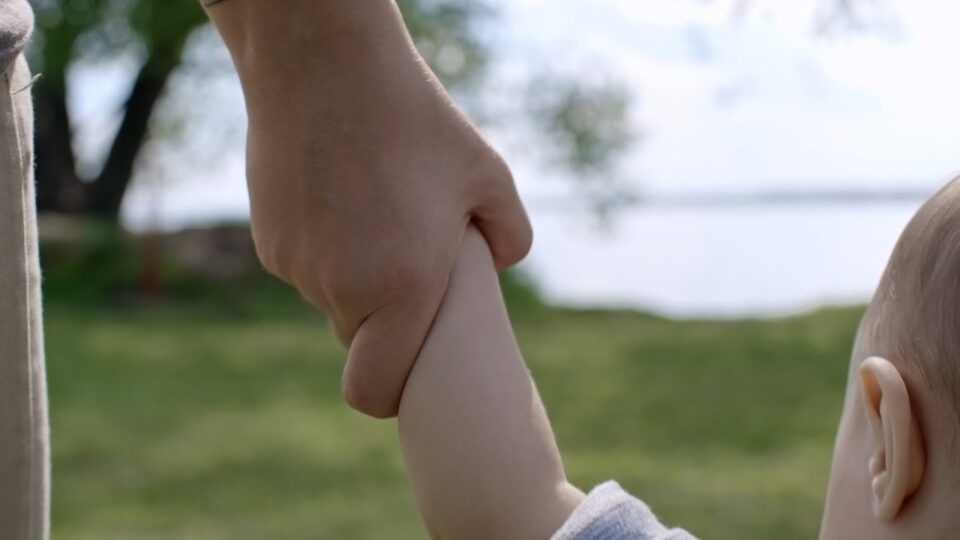 baby holding an adult's hand with a lake in the background