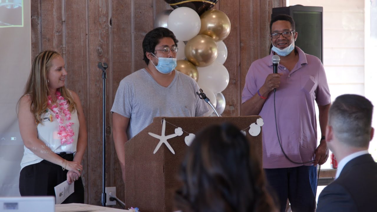 a person with a mask on stands a t podium with two other people