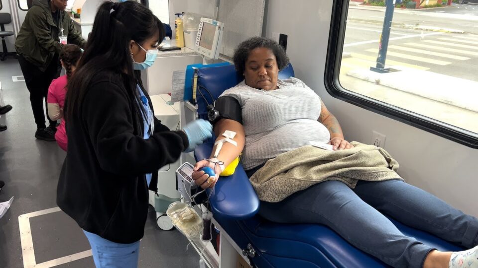 person sits in reclined chair donating blood as a nurse stands beside the chair