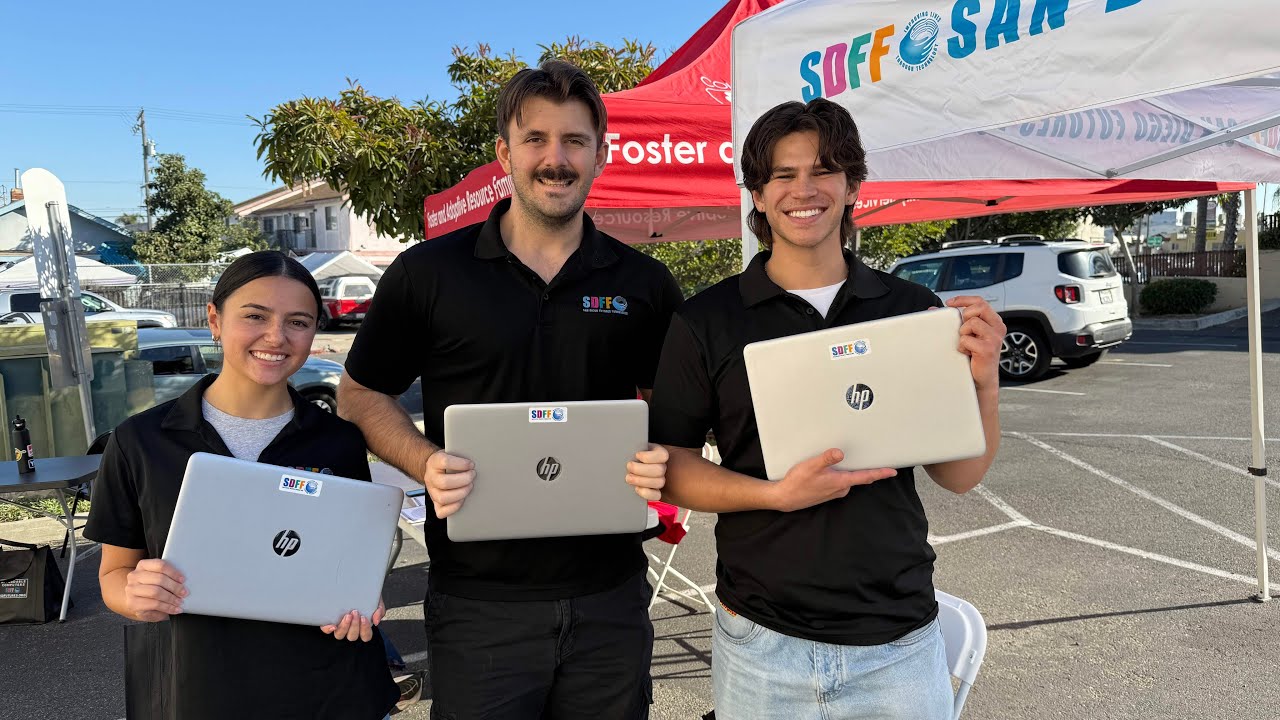three hold laptops next to pop-up shade canopies in a parking lot