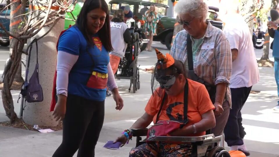 person wearing superhero shirt stand next to person in wheelchair wearing mask