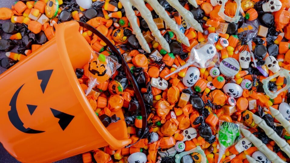 a pail decorated like a pumpkin spills candy out with two skeleton hands holding the candy
