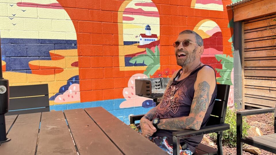 Man wearing sleeveless shirt sits at an outdoor table with a mural and grill behind him