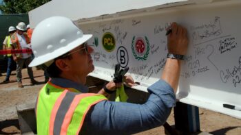 Chair Fletcher signs a large beam while wearing a hard hat