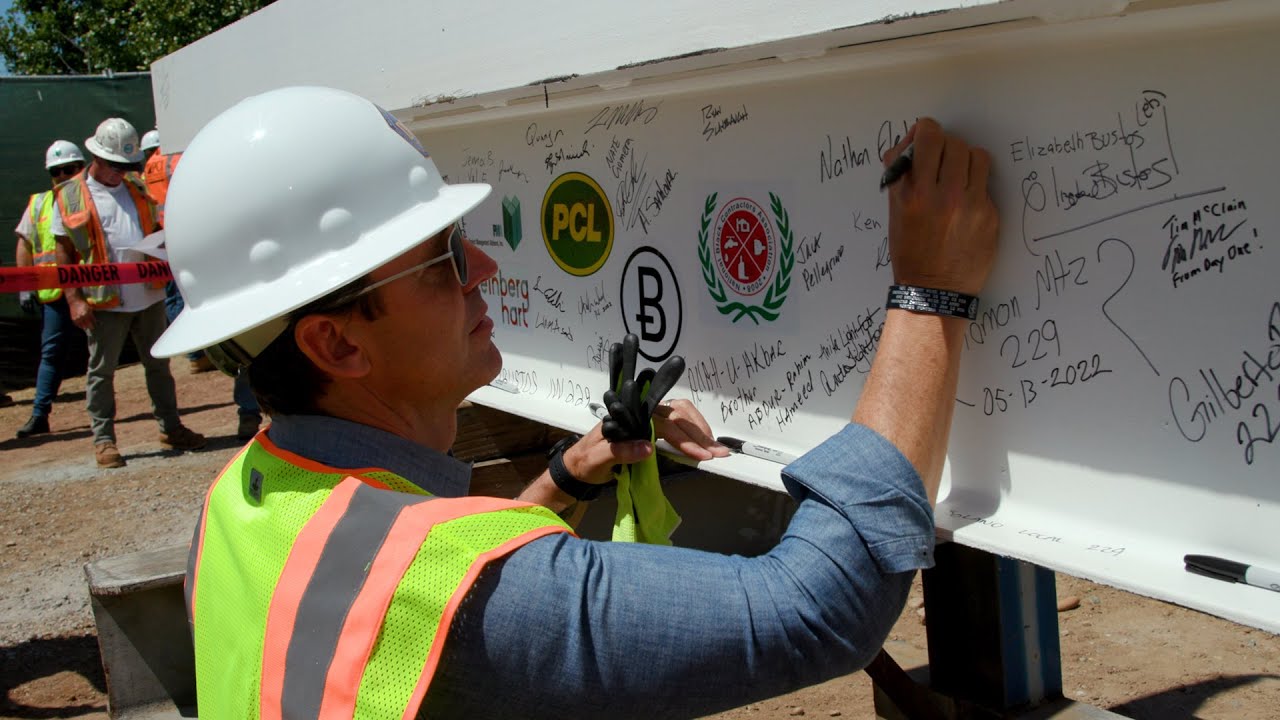 Chair Fletcher signs a large beam while wearing a hard hat