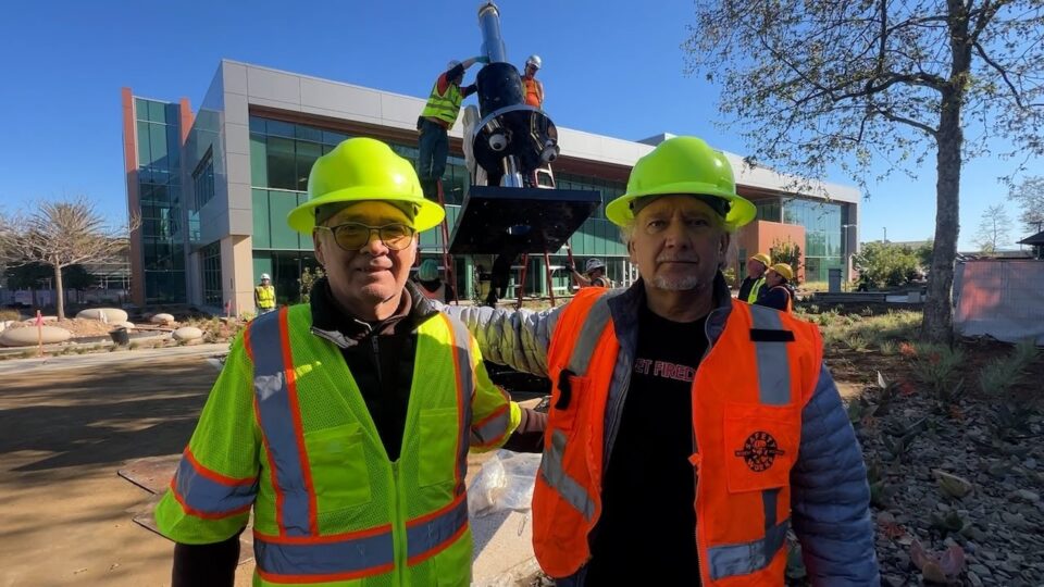 2 people wearing construction hats and vests stand in front of sculpture shaped like a microscope