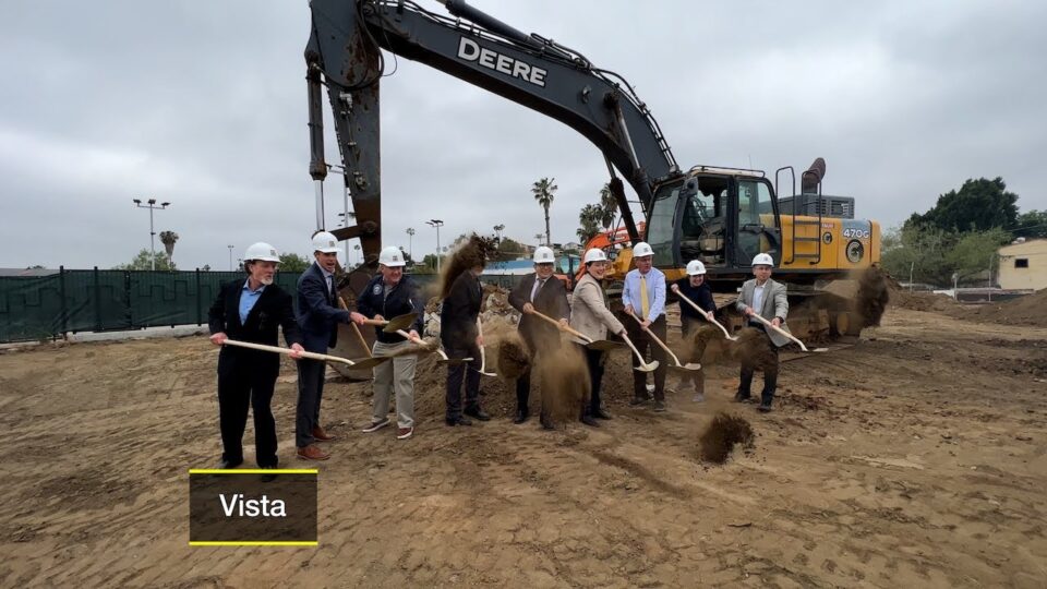 group with hard hats shovels dirt in front of construction equipment
