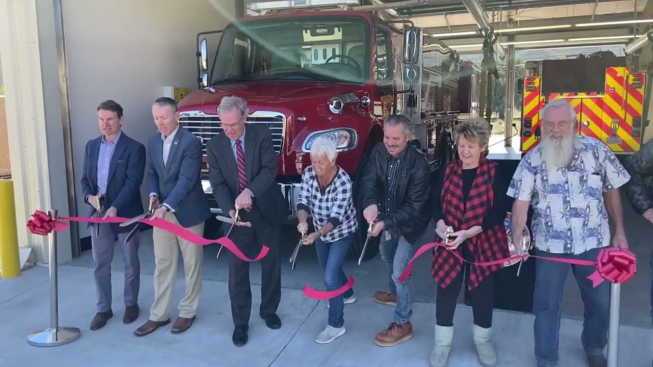 7 people cutting a ribbon in front of a fire station